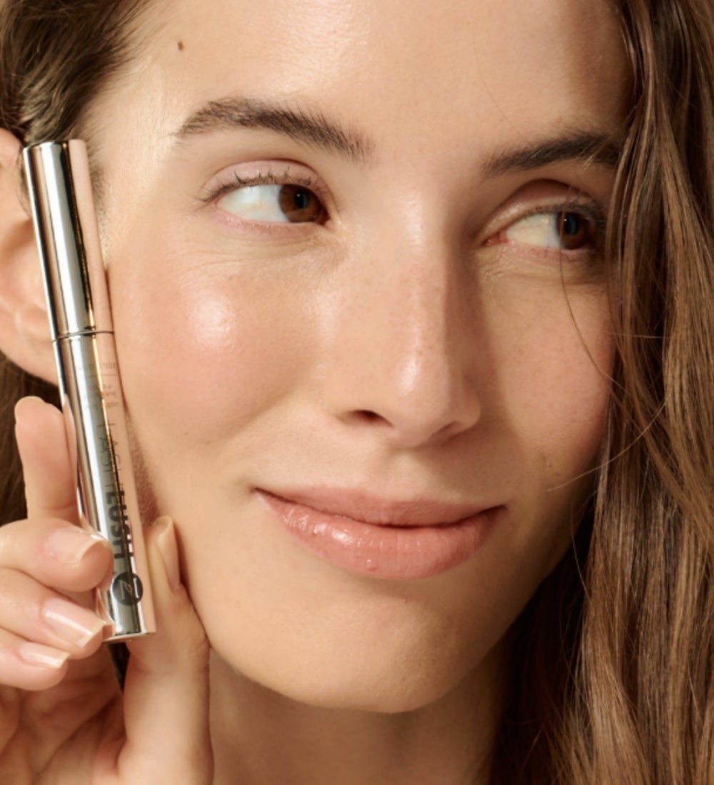 A woman holding a LashLush Serum tube beside her face.
